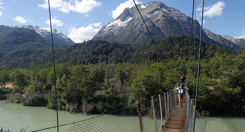 A group of people make their way over a swing bridge that hangs over a river. In the background, there are thick green trees in front of a mountain. 